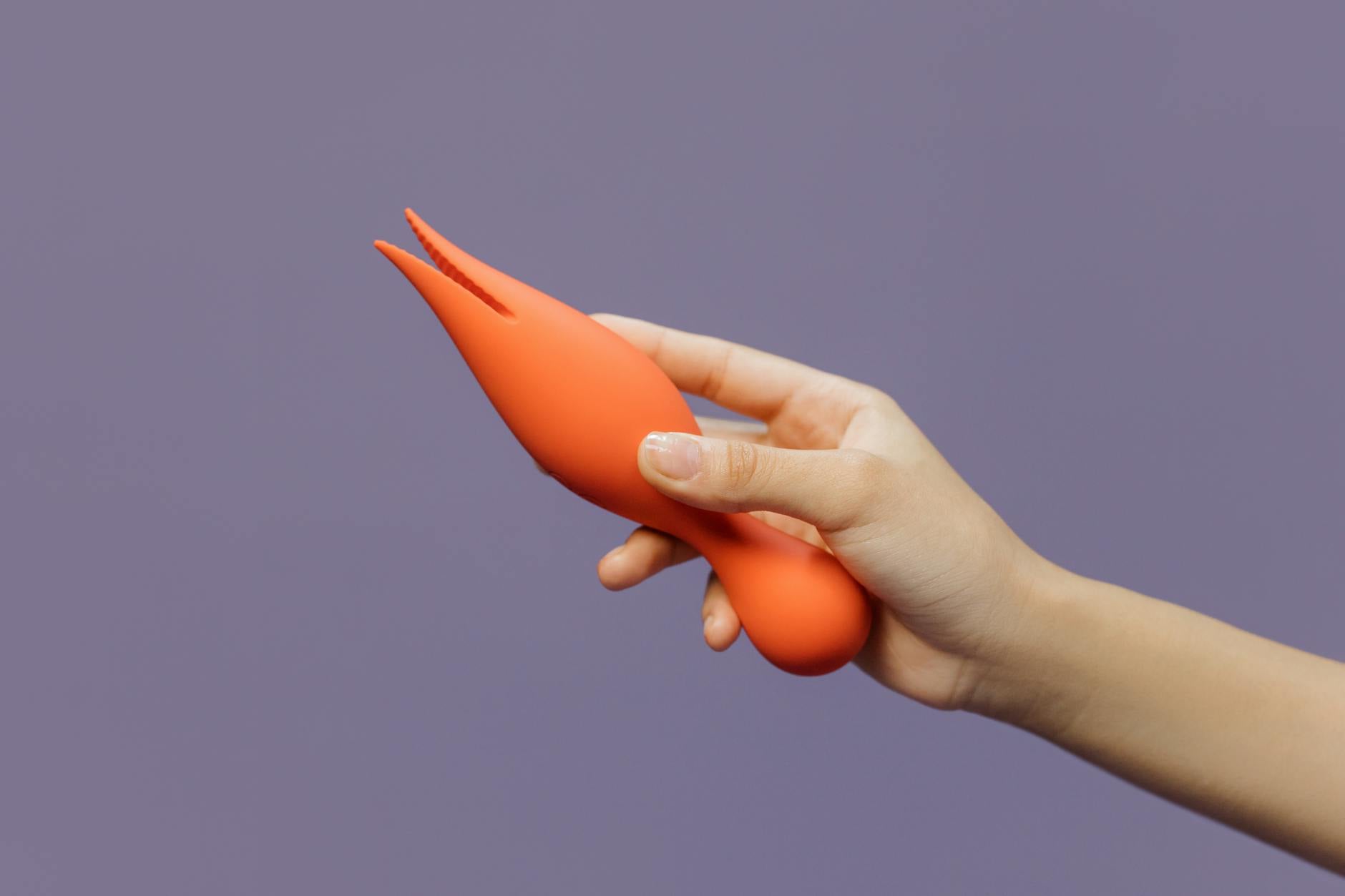 Close-up of a hand holding an orange lemon vibrator against a minimalistic purple backdrop.
