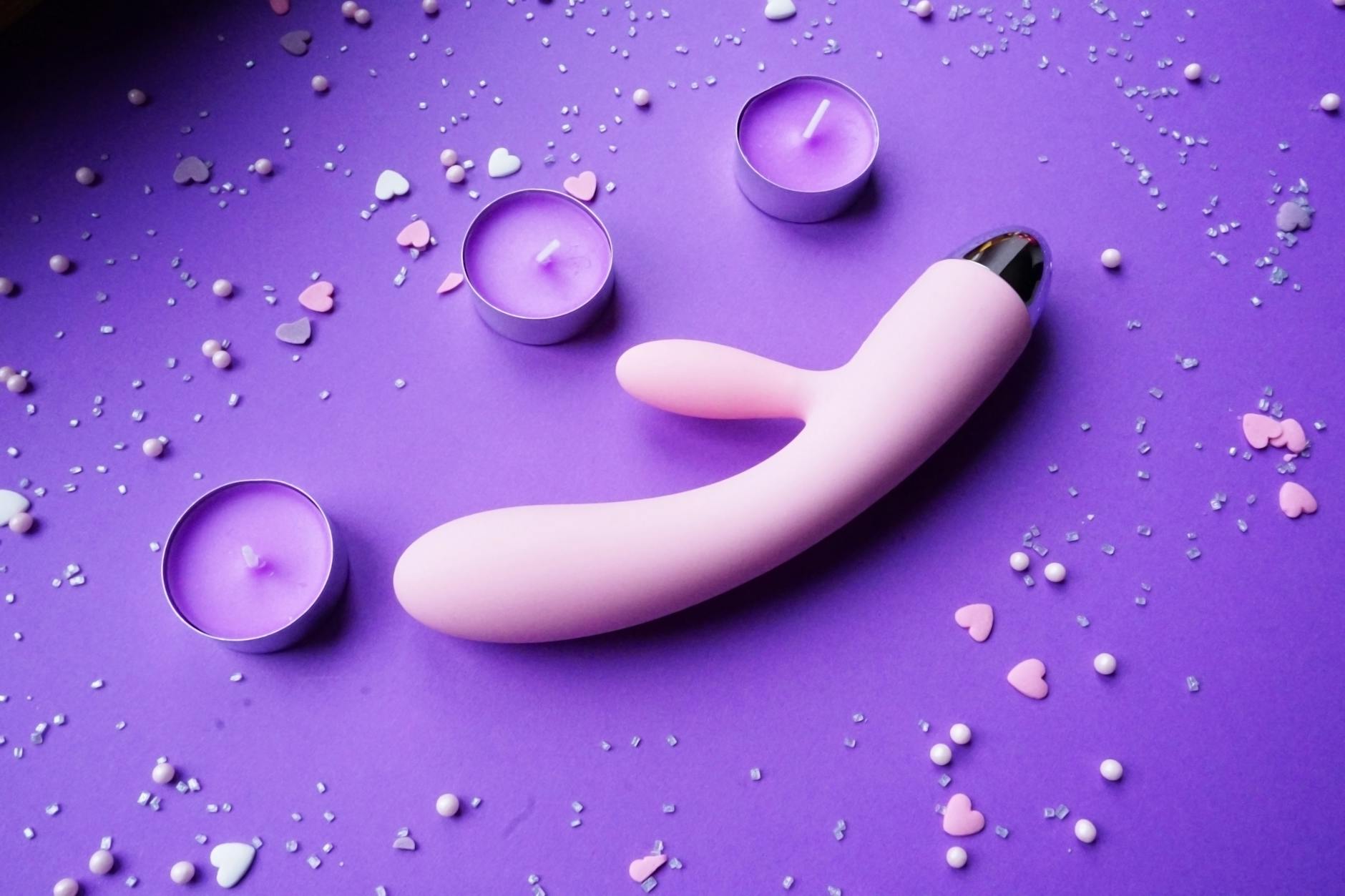Pink vibrator arranged on a purple background with heart confetti and candles for a romantic atmosphere.